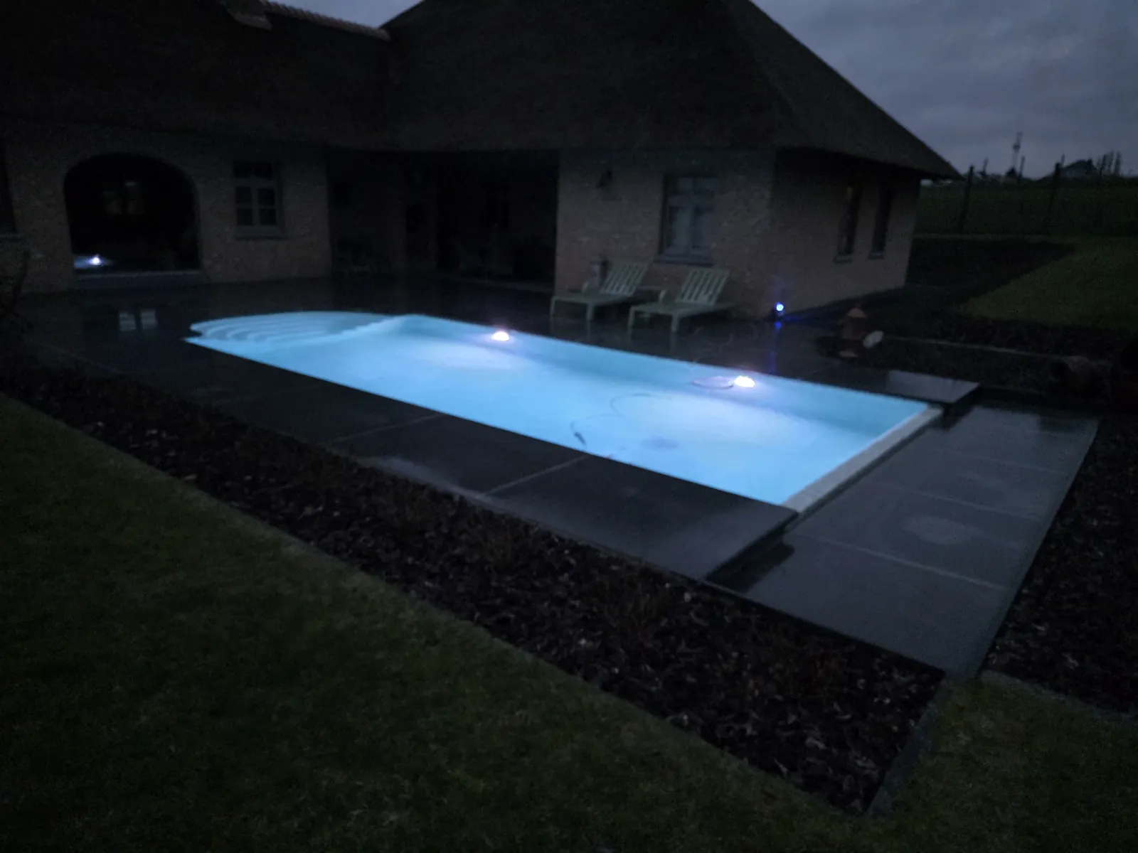 Verlicht zwembad met een overlooprand bij een woning in de avond